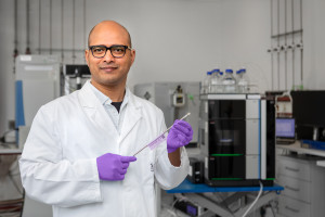 Dr Laxmikanth Kollipara stands in the laboratory holding a silver-coloured column. In the background are HPLC systems.