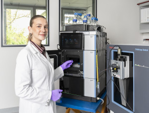 Neele Rottmann is wearing a white lab coat and purple gloves and is operating a mass spectrometry analyser in a laboratory. Next to her is a liquid chromatography system connected to a mass spectrometer via several tubes. There are numerous sample vials on the instrument. As she places a sample into the system, she looks into the camera.