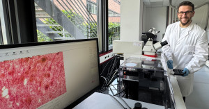 Kevin Hau trägt einen weißen Kittel mit ISAS-Logo. Er steht in einem Labor an einem Mikroskop. In der linken unteren Ecke des Bildes ist ein Computerbildschirm zu sehen. Auf dem Monitor ist ein mikroskopisches Bild von rötlichem Gewebe geöffnet.
